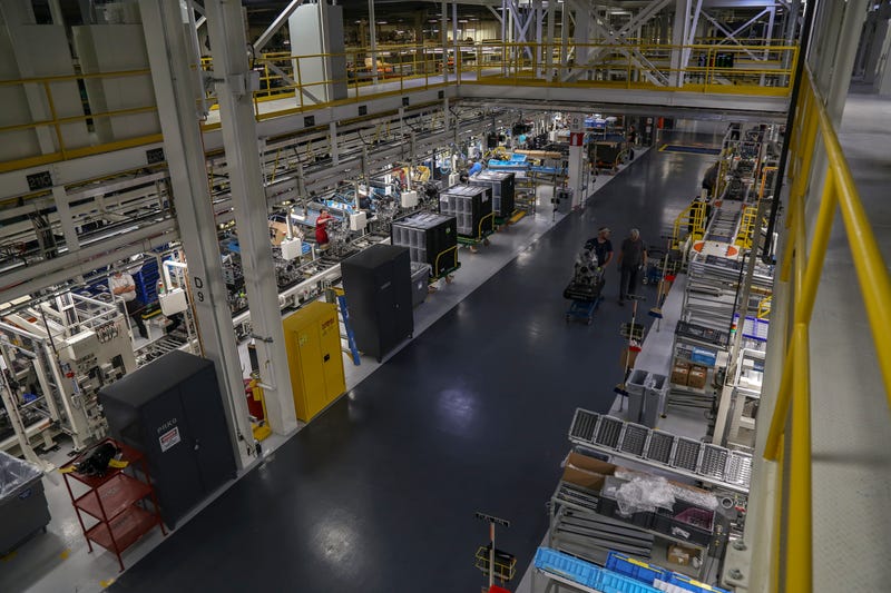 Aerial view of a busy factory floor with workers, machinery, and conveyor belts under bright lighting. The space feels organized and industrious.