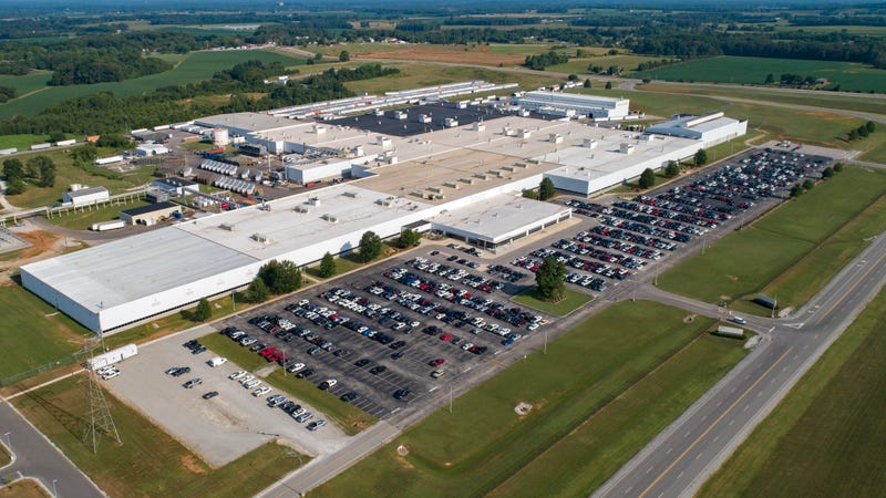 Aerial view of a large industrial complex with multiple white-roofed buildings and parking lots, surrounded by green fields and roads under a clear blue sky.