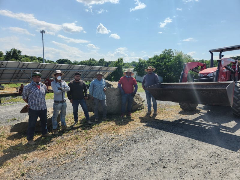 Farmers stand by a row of solar panels and a bulldozer. Hepworth is renowned for it's green energy and fair labor practices.
