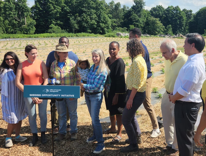 A cute baby with a hat is joined by farm workers, state politicians and OCM staff at a press conference on the future of cannabis agriculture in New York. Gail Hepworth (in the blue flannel) and Amy Hepworth (in the yellow flannel) stand in the center.