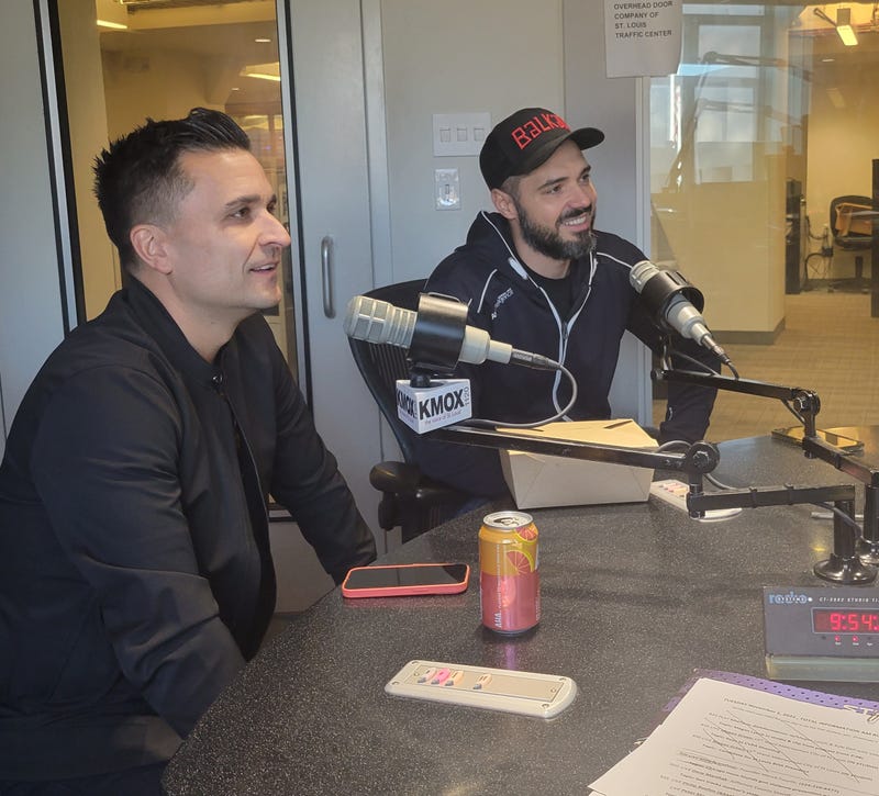 two men sit at kmox microphones they're both wearing black jackets and one wears a black hat that says balkan
