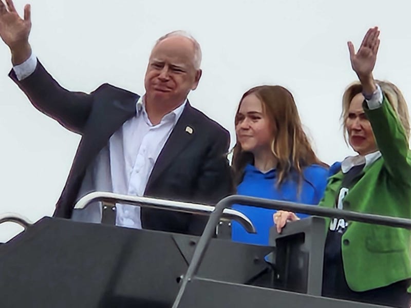 Minnesota Governor and Vice Presidential Candidate Tim Walz waves at onlookers as he boards a plane at MSP Airport with daughter Hope and wife Gwen.