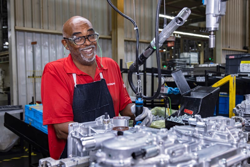 Smiling worker in a red shirt and apron operates machinery on an industrial assembly line, conveying a positive and focused work environment.