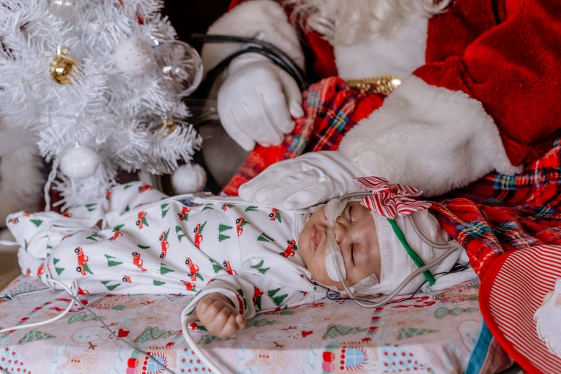 Tiny baby in onesie decorated with red truck carrying Christmas trees