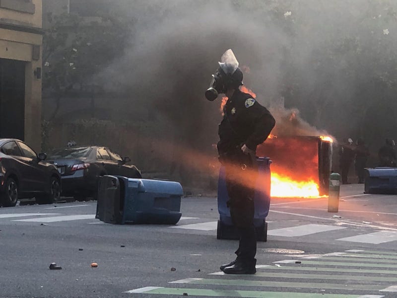 An officer stands alone as a dumpster burns in the background.