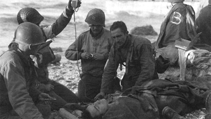 Medics on D-Day beach head in Normandy treat one of their own with an IV as the seawater rolls in behind them on June 6, 1944.