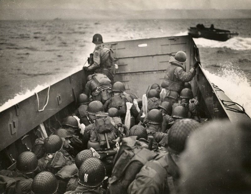 Tightly packed troops crouch inside their LCVPs (Landing Craft Vehicle, Personnel), as it plows through a wave. In the distance is the coast of Normandy.