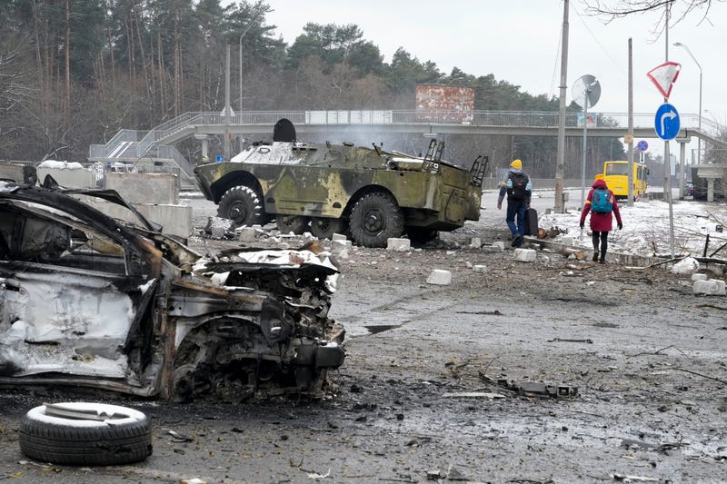 People walk by a damaged vehicle and an armored car at a checkpoint in Brovary, outside Kyiv, Ukraine, Tuesday, March 1, 2022