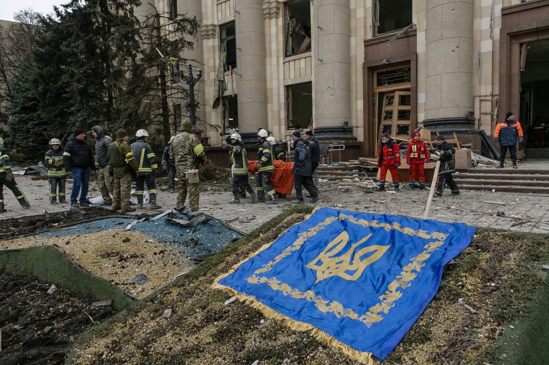 Ukrainian emergency service personnel walk past a Ukrainian coat of arms while carrying a body of a victim following shelling of the City Hall building in Kharkiv, Ukraine, Tuesday, March 1, 2022
