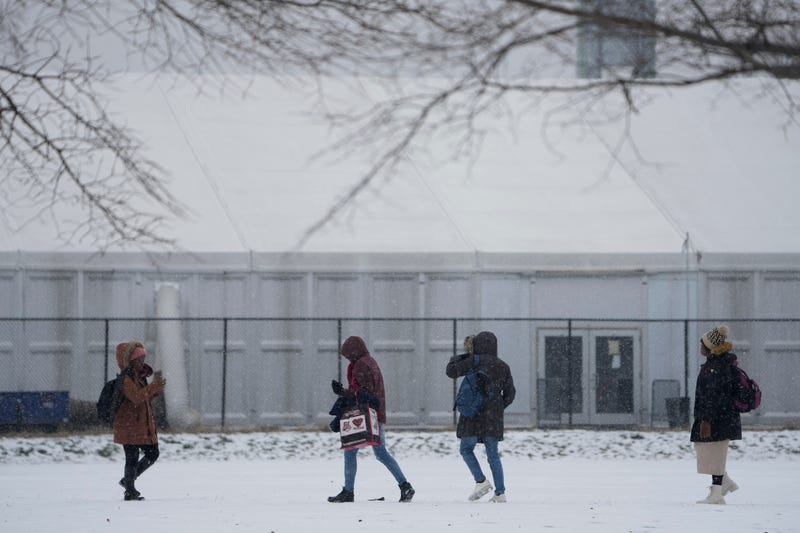 People walks across a snowy athletic field in front of a temporary shelter for migrants on Randall's Island in New York, Friday, Jan. 19, 2024