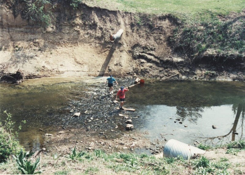 An undated photo from the 1980s, of two teenagers stepping on rocks and wooden planks on Coldwater Creek. The photo is from a scrapbook kept by Sandy Delcoure, who lived on Willow Creek in Florissant and donated the scrapbook to the Kay Drey Mallinckrodt Collection.