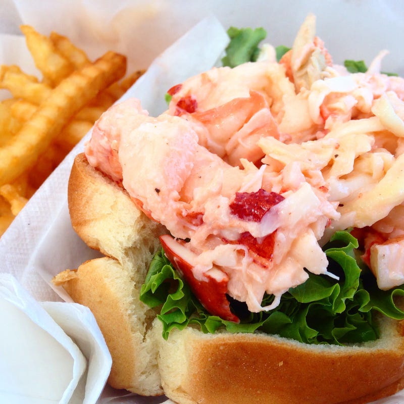 A lobster roll loaded with chunks of lobster meat on a bed of lettuce, with fries on the side nearby.