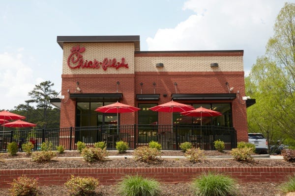 Exterior of a Chick-fil-A restaurant. Tall building with a storefront and a dining room. There's also an outside dining area with tables and umbrellas. Landscaping and a low brick wall separate the restaurant from the sidewalk.