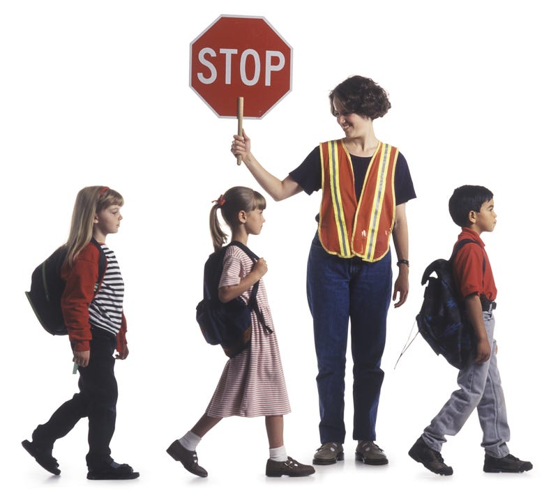 A group of children walk in a line while assisted by a crossing guard