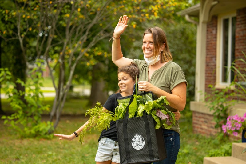 Mom and daughter waving while holding a Market Wagon reusable tote overflowing with vegetables