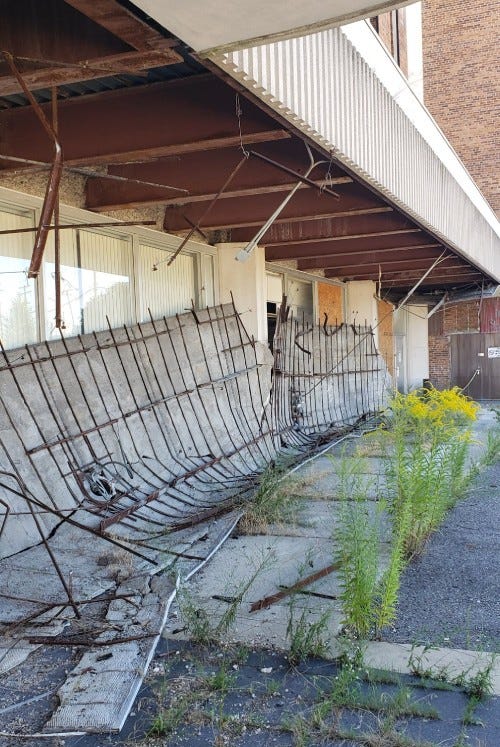 Broken down fencing leans against the lower floor of the office building. The parking lot next to it is broken up, uneven and overgrown with weeds.