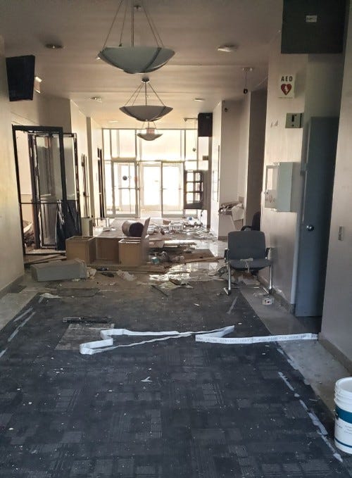 Inside of the office building lobby. The carpet is covered in crumbling drywall. A flipped over desk, trash can, chair and bucket can be seen on the floor along with a lot of trash.