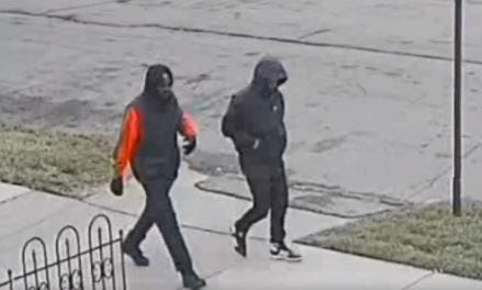 Surveillance footage of two men walking along a sidewalk in a neighborhood. Both wear black pants. One has a red and black jacket. The other has a black jacket. Both have their hoods up. They are alleged carjackers.