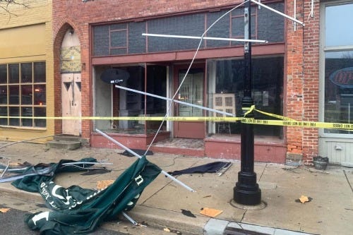 An awning and signage from a storefront hang from the roof of a building. Glass and wood debris are scattered on the ground.