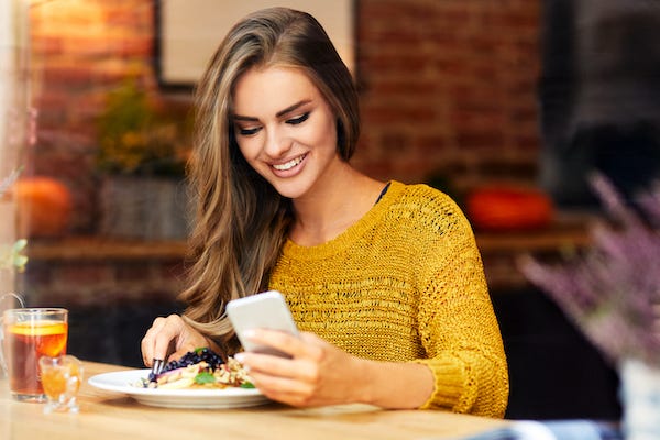 Beautiful smiling young woman looking at phone while sitting and eating in a cafe - stock