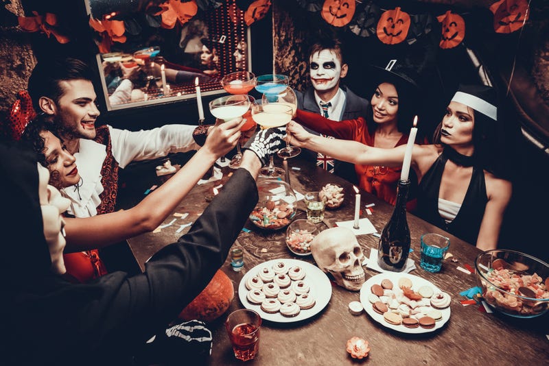Group of Young Happy People Wearing Costumes at Halloween Party Sitting at Table and Drinking Cocktails.