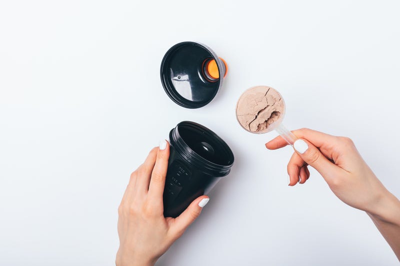 Woman's hands putting scoop of chocolate protein powder into black plastic shaker to prepare sports drink for muscle growth, top view on white table.