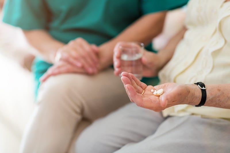 stock photo. elderly woman holds a glass of water in one hand and pills in another. a healthcare worker sits beside her.