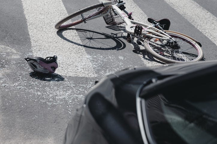 A bicycle lies on the ground at a crosswalk. A helmet lies next to it. The front of a black vehicle can be seen.