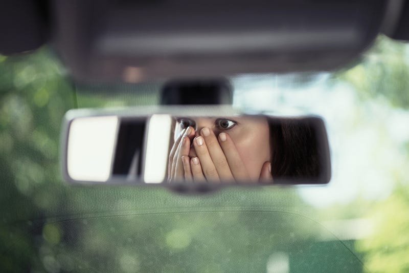 Reflection of young woman with frightened eyes covering her face with hands in the car rear view mirror.