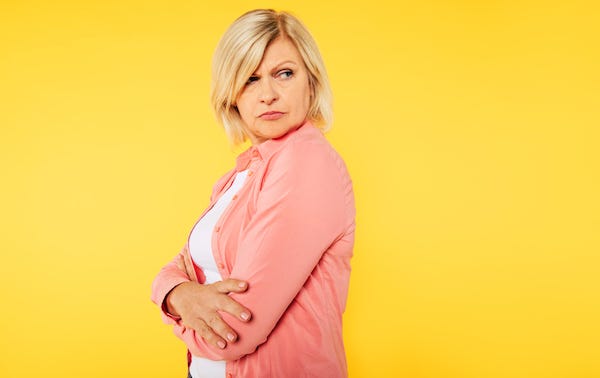 Angry blonde senior woman with crossed arms is posing over yellow background