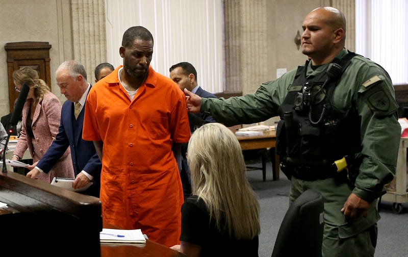 R. Kelly stands in a courtroom in a orange jumpsuit, while a guard escorts him out