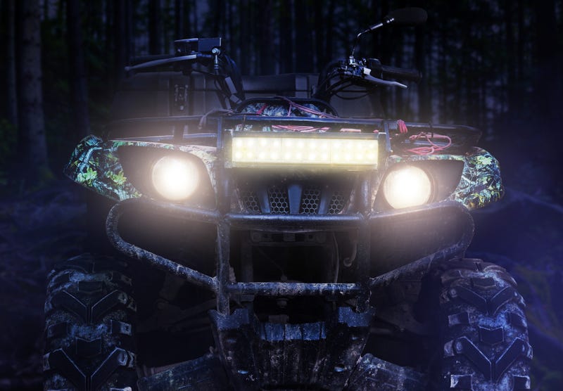 a camouflaged dirty offroad hunting atv vehicle standing at night in forest with headlights turned on, front view.