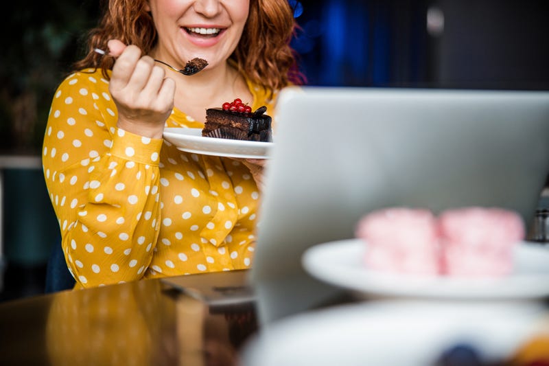 Cheerful woman eating cake and using laptop