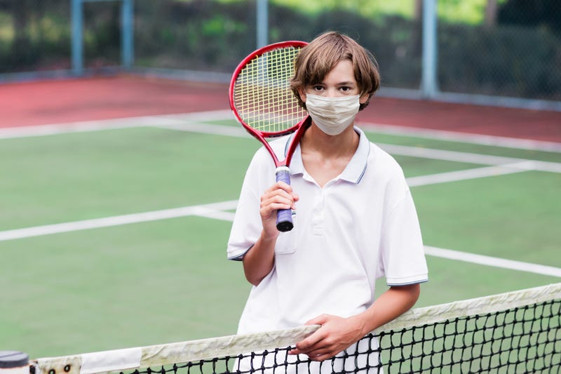 Boy in face mask playing tennis on outdoor court during coronavirus outbreak.