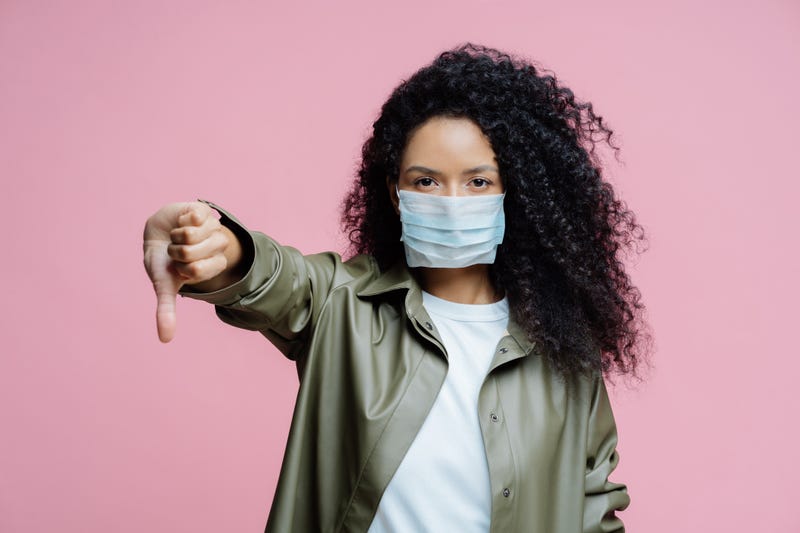 Image of serious curly haired young woman wears medical mask, keeps thumb down, shows dislike gesture, avoids spreading of corona virus, cares about health.