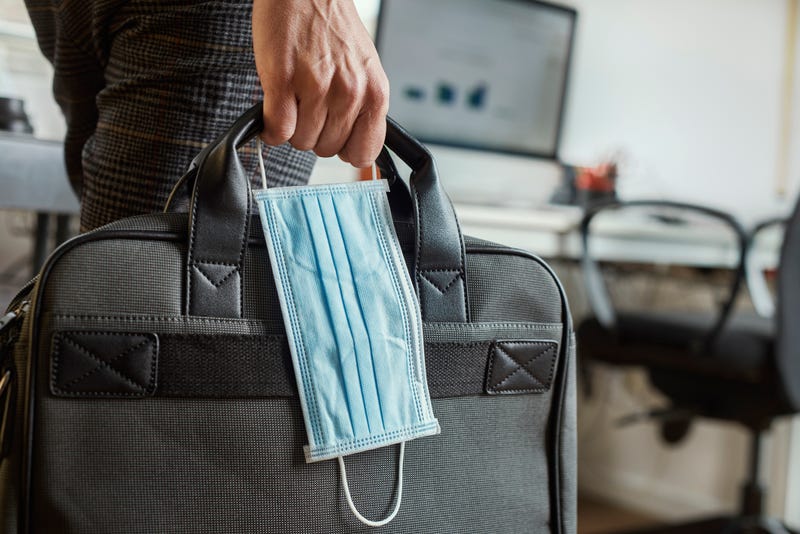 A closeup of a young man in an office holding a briefcase and a surgical mask in his hand.