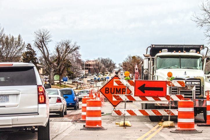 A city road under construction. Traffic barrels and other signage block several lanes. Work trucks are onsite, and drivers shift into the only open lane.