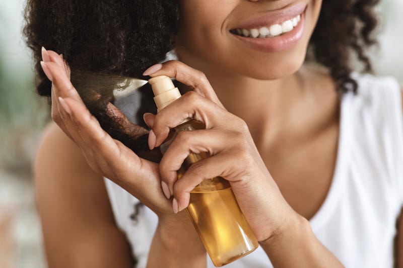 Split Ends Repair Treatment. Smiling African Woman Applying Essential Oil Spray On Her Curly Brown Hair At Home