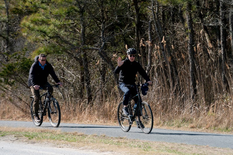 US President Joe Biden (R) waves while riding his bike through Cape Henlopen State Park in Rehoboth Beach, Delaware, on March 20, 2022. 