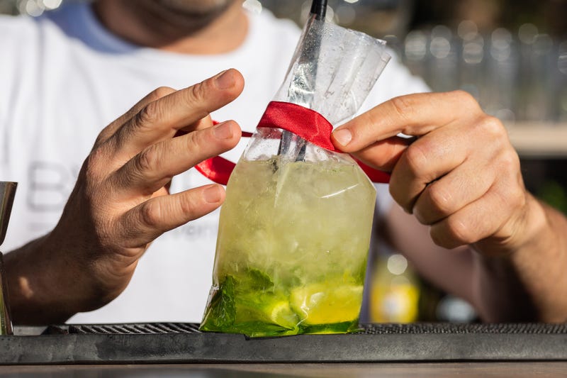 Two bartender's hands tie a red ribbon to a plastic take-out can containing a lime, lemon and mint mojito. Above a bar (pub) outside, it's daytime.
