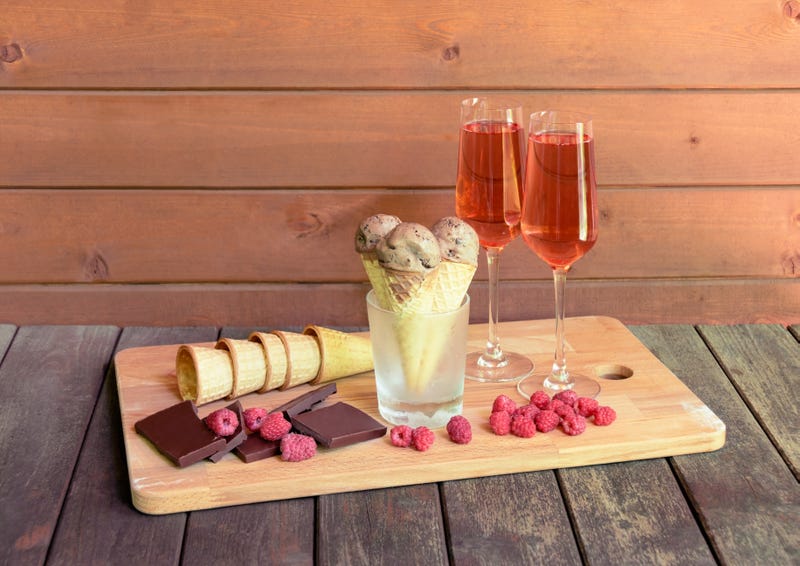 Glasses of rose champagne, waffle cones with chocolate ice cream in glass as stand, pieces of dark chocolate, fresh raspberry and empty waffle cones near on rustic wooden table.