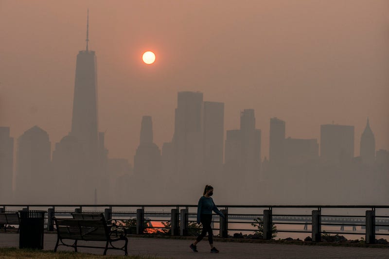 A woman walks as the sun rises behind the One World Trade Center and the New York skyline, while the smoke from Canada wildfires covers the Manhattan borough as it is seen from the Liberty State Park on June 8, 2023 in New Jersey
