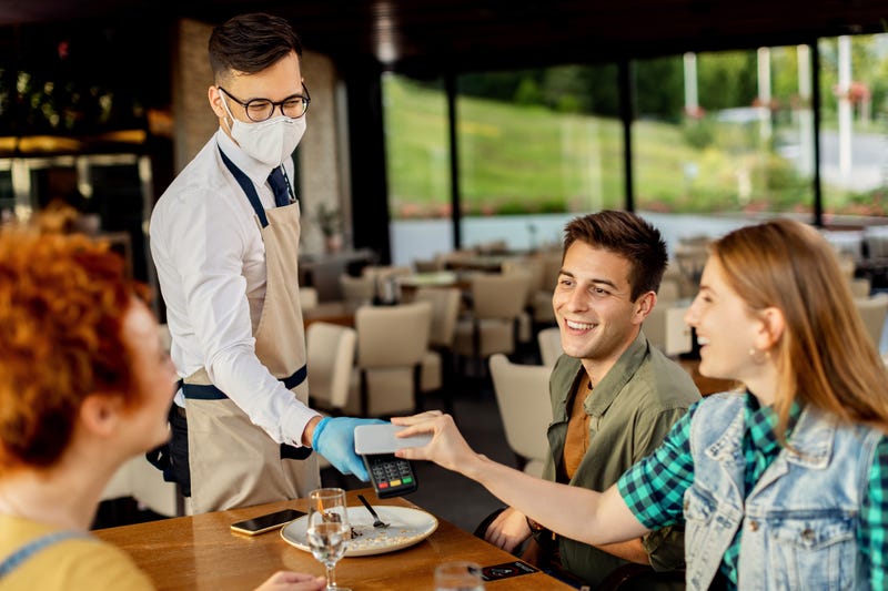 Group of friends paying contactless with mobile phone to a waiter in a cafe. Focus is on waiter.