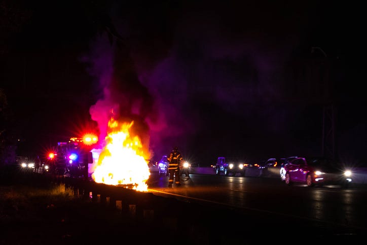 A car on fire on a highway at night. Police are behind the wreck. Firefighters stand to the side, working to douse the fire. Other cars pass by on the side.