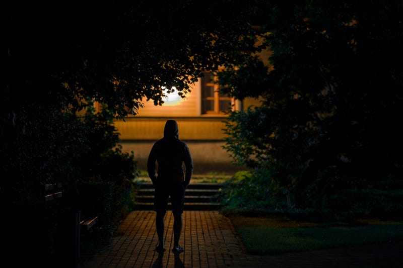 Hooded man standing on sidewalk at dark park in summer night.