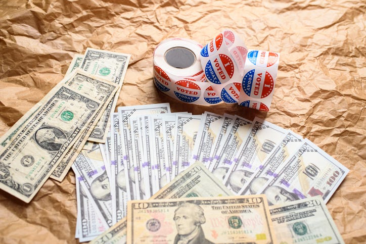 A stack of hundred dollar bills is fanned out on a table next to a roll of stickers that read "I voted."