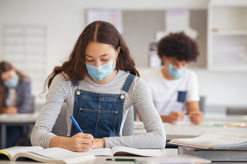 High school student taking notes from book while wearing face mask.