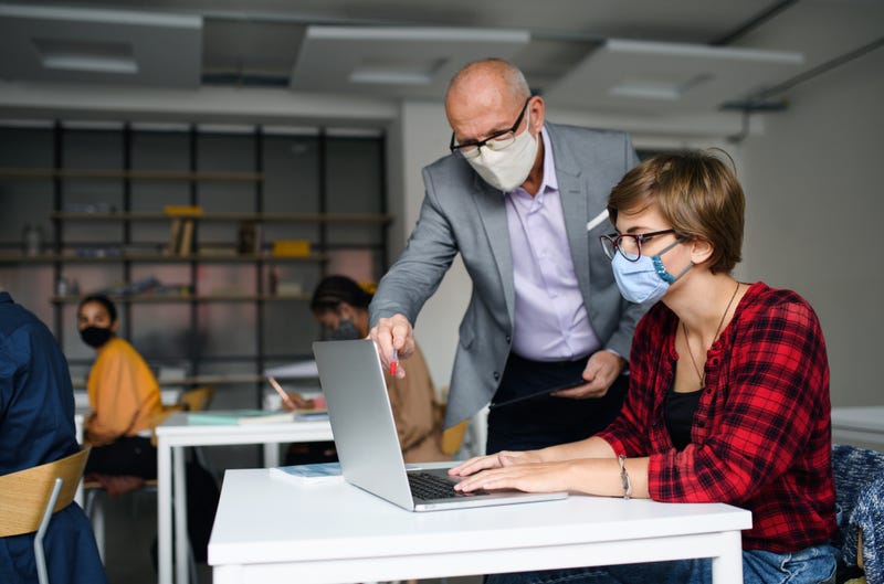 File photo of a masked teacher helping a masked student at her desk.