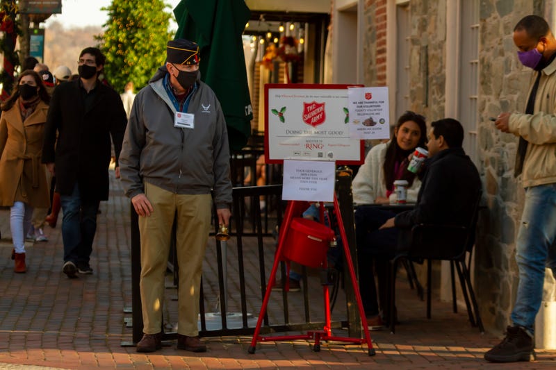 An elderly veteran with an airman side cap hat and face mask is standing next to a Salvation Army donation bucket, as a volunteer bell ringer.