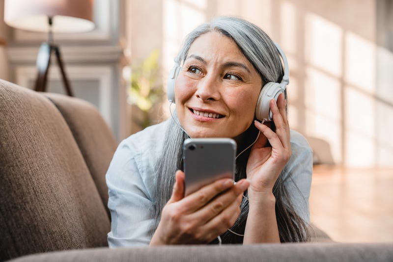 Woman lying belly down on couch holding a cellphone and enjoying audio on her headphones.